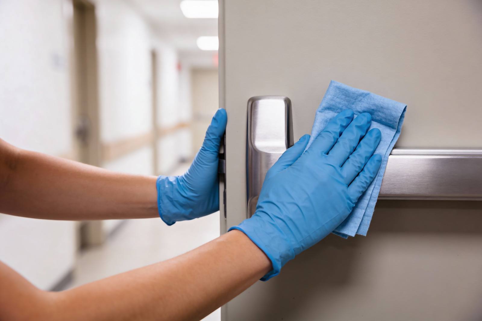 Gloved hand disinfecting a door handle and push plate in a clinic corridor