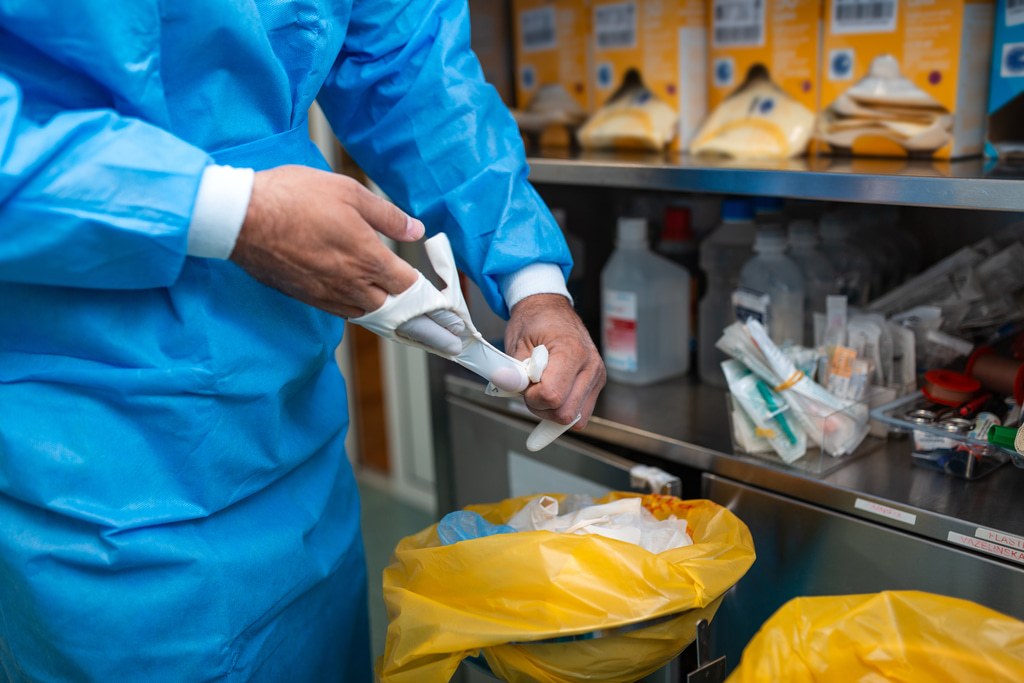 Cleaner in a gown removing gloves beside a clinical waste bin